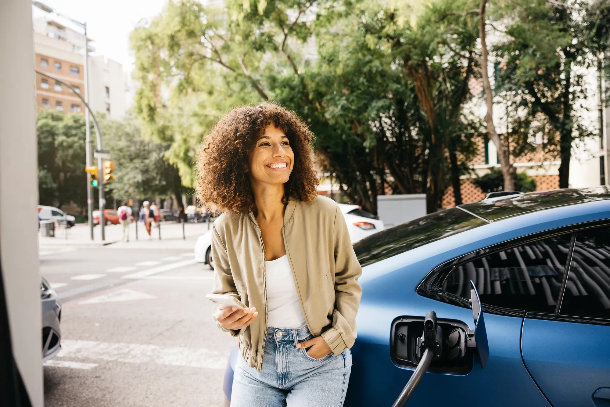 Smiling person standing next to their electric vehicle while it charges in a city setting